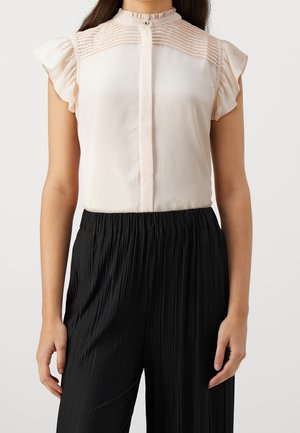 Vrouw draagt een lichtroze mouwloze blouse met geruite schouders en geplooide zwarte broek met hoge taille tegen een witte achtergrond.