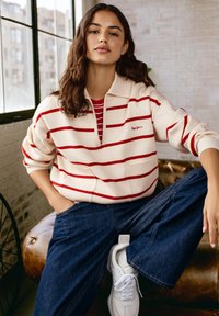 Pullover crema con strisce rosse, colletto con zip, tasca frontale. Indossato sopra una maglietta a strisce rossa e bianca, con jeans larghi blu e sneaker bianche.