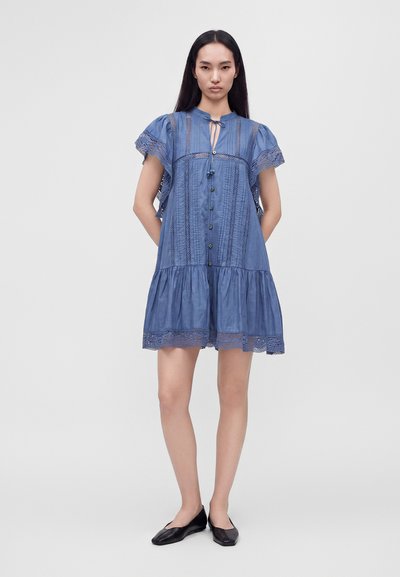 Jeune femme debout les mains dans le dos, portant une courte robe bleue avec des boutons et des détails en dentelle, associée à des chaussures plates noires.