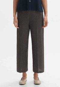 Kobieta nosząca szerokie spodnie z geometrycznym wzorem w kształcie diamentu w ciemnych i jasnobrązowych odcieniach, w połączeniu z beżowymi płaskimi butami.