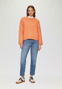 Pomarańczowy sweter ribbed, luźny krój, opadające ramiona, w zestawieniu z niebieskimi dżinsami o prostym kroju i wielokolorowymi sneakersami z czarnymi akcentami.