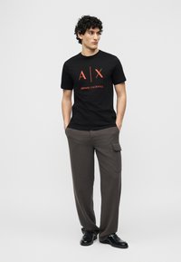 Νέος άνδρας με σγουρά μαλλιά, φορώντας μαύρο T-shirt Armani Exchange, γκρι παντελόνι cargo και μαύρα παπούτσια, στέκεται με τα χέρια στις τσέπες.