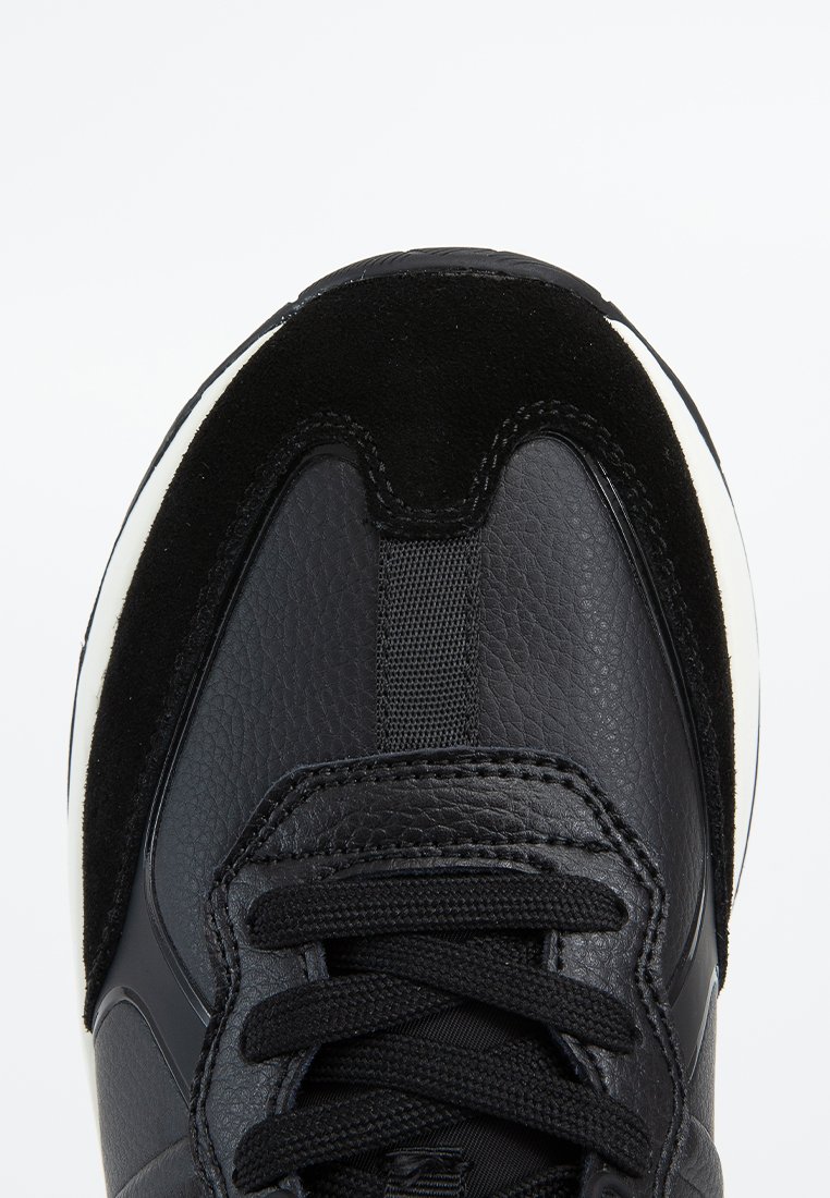 Chaussure de sport noire avec une tige en cuir lisse et des accents en daim, languette en mesh texturé et lacets noirs. Détail de semelle intercalaire blanche.