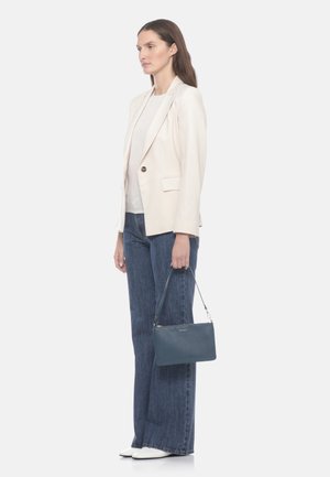 Donna che indossa un blazer beige chiaro, camicia bianca, jeans blu a gamba larga, scarpe bianche, che tiene una borsa blu per la tracolla al suo fianco.