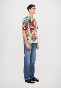 Cămașă cu mânecă scurtă, multicoloră, cu un amestec de modele, asortată cu blugi ușor albaștri și sandale negre.
