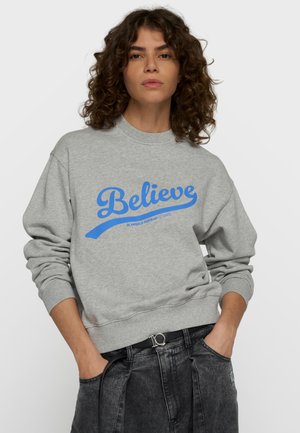 Frau mit lockigem Haar, die einen grauen Pullover mit blauem "Believe" Text trägt und die Hände in den Taschen der schwarzen Jeans hat.