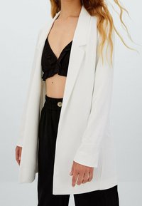 Femme portant un blazer blanc sur un crop top noir à volants et un pantalon noir taille haute avec un détail à boutons.