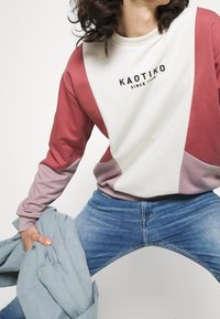Sweatshirt color block in morbido cotone con pannelli rosa, bordeaux e crema. Include un collo a giro e un logo grafico. Indossato con jeans in denim.