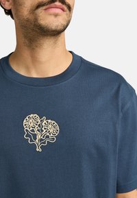 Navyblaue Baumwoll-T-Shirt mit Rundhalsausschnitt, das ein gesticktes Herzdesign mit floralen Elementen in Cremefarbe auf der Brust zeigt.