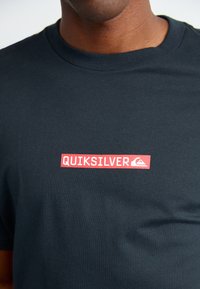 Homme portant un t-shirt noir avec un logo rectangulaire rouge affichant "QUIKSILVER" et une icône de montagne ondulée sur la poitrine.
