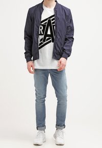 Marinblå bomberjacka med ribbat krage, grå grafisk t-shirt, ljusblå slim-fit jeans och vita sneakers. Avslappnad urban outfit.