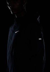Czarna kurtka Nike z refleksyjnym logo na klatce piersiowej i małym refleksyjnym akcentem na ramieniu; ma wysoki kołnierz i gładką teksturę.