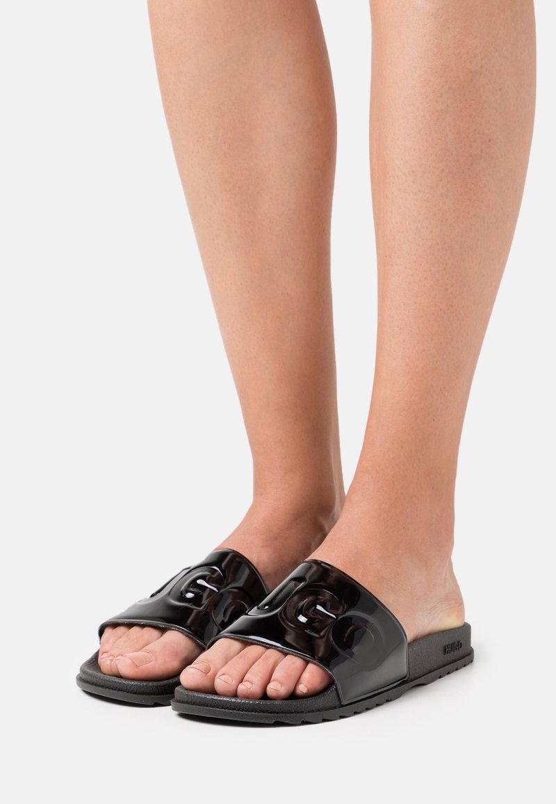 Barfötter i svarta glänsande slip-on sandaler med upphöjd "HUGO" logotyp på remmarna mot en enkel vit bakgrund.