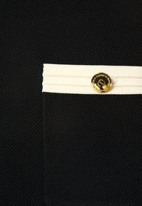 Tissu noir texturé avec une bordure blanche côtelée et un bouton doré gravé de texte sur une poche de vêtement.