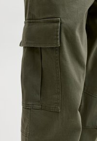 Gros plan sur la poche d'un pantalon cargo vert olive avec rabat et couture visible sur la texture du tissu.