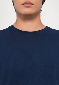 Gros plan d'une personne portant un t-shirt uni bleu foncé à col rond, visible du cou jusqu'au milieu de la poitrine sur un fond neutre.