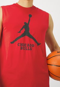 Rød ærmeløs t-shirt med et sort Jumpman-logo og teksten "CHICAGO BULLS" nedenfor. Glat stof, atletisk fit, holdt tæt på en basketball.