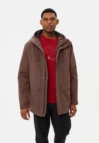 Giacca impermeabile marrone con zip frontale, due tasche anteriori e cappuccio foderato grigio, indossata sopra una t-shirt grafica rossa e pantaloni neri.