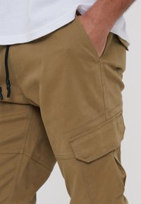 Pantalons cargo en khaki avec une texture lisse, dotés de poches latérales et d'une taille à cordon réglable. Le tissu semble robuste et durable.