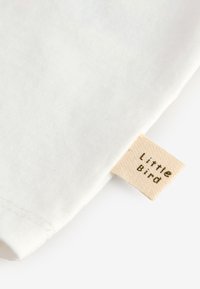 Fehér pamutanyag sima textúrával, egy kis bézs címkével, amelyen zöld nyomtatással a "Little Bird" felirat olvasható.