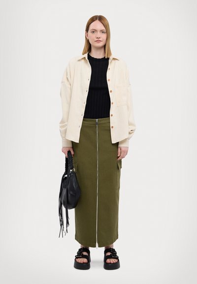 Camisa de botones color blanco roto, suéter negro de canalé, falda verde olivo con cremalleras y bolsillos, y sandalias de plataforma negras con detalles trenzados.