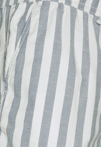 Vitt tyg med grå vertikala ränder. Har ett elastiskt midjeband och en avslappnad passform, med strukturerat bomullsmaterial. Ingen synlig hårdvara.