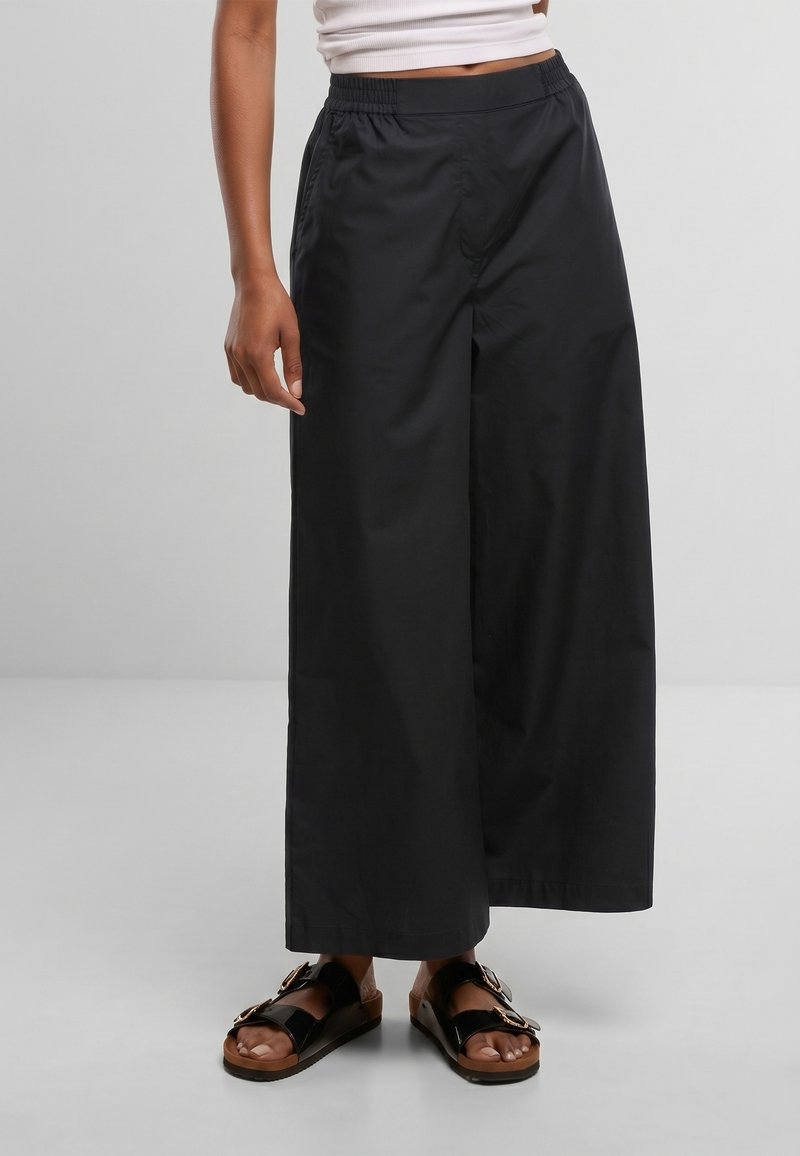 Personne portant un pantalon noir à jambes larges, un haut court blanc et des sandales noires à boucles, debout sur un fond gris.