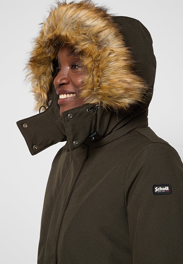 Parka isolante vert olive avec une capuche doublée en fausse fourrure, fermetures éclair et à pression, et un patch logo noir sur la manche gauche.