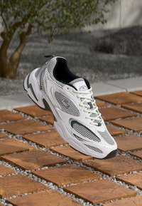 White athletic shoes with mesh upper, black and green accents, padded collar, and cushioned sole, positioned above textured tiles.