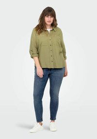 Olive zelená košeľa na gombíky s dvoma náprsníkovými vreckami, vyhrnutými rukávmi, v kombinácii s denimovými skinny džínami a bielymi teniskami. Hladká textúra po celej ploche.