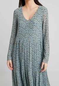 Sukienka w kwiaty w jasnym niebieskim kolorze z czarnymi i pomarańczowymi wzorami. Ma długie rękawy, dekolt w serek oraz warstwową, zwiewną spódnicę. Miękka tekstura materiału.