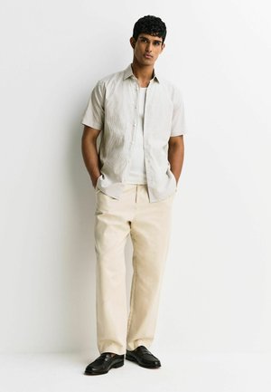Man standing with hands in pockets wearing light short-sleeve button-up shirt, white undershirt, beige pants, and black shoes against white background.