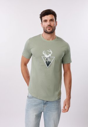 Homme portant un t-shirt vert clair avec une tête de cerf blanche et une forêt à l'intérieur d'un triangle, associé à un jean bleu clair, debout devant un fond uni.