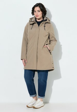 Vrouw staand, gekleed in een beige capuchonjas, omgeslagen blauwe jeans en witte sneakers tegen een effen lichte achtergrond.