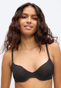 Νεαρή γυναίκα με κυματιστά μαλλιά που φοράει μαύρο σουτιέν με μπανέλα και λεπτές τιράντες, σε απλό ανοιχτό φόντο.