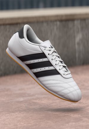 Biały sneaker z czarnymi paskami i gumową podeszwą, sznurowany, unosi się nad różowawą betonową powierzchnią z rozmytym tłem.