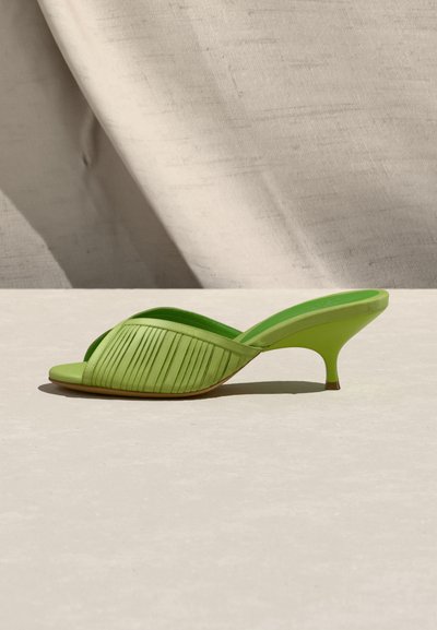 Ανοιχτό πράσινο mule με πτυχωτό λουράκι και χαμηλό τακούνι στιλέτο, τοποθετημένο σε ουδέτερη επιφάνεια με υφασμάτινο φόντο.