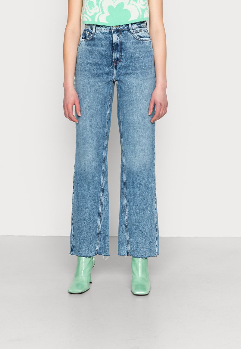 Jeans de boca larga azul claro com cintura alta, textura desbotada e bainha crua. Combinados com botas de tornozelo em couro envernizado verde.