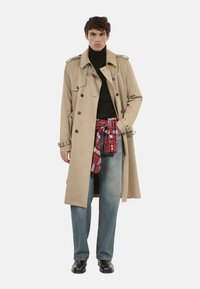 Gabardina beige con diseño de doble botonadura, epaulettes en los hombros y cuello alto negro. Combinada con jeans azules y una camisa de cuadros rojos atada a la cintura.