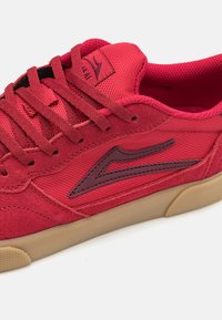 Punaiset tennarit, jotka on valmistettu tekstuurikankaasta ja mokasta, sisältäen tummanpunaisen logoversion, litteät nauhat ja vaaleanruskean kumipohjan.