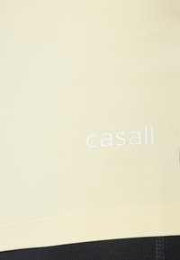 Svetložltý športový top s hladkou textúrou. Na prednej strane sa nachádza biele logo "casall". Navrhnutý pre pohodlie a pohyb.