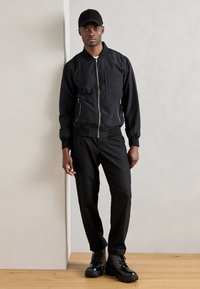 Schwarze Bomberjacke mit Reißverschluss, gerippten Bündchen und seitlichen Taschen; kombiniert mit schwarzen Hosen und klobigen schwarzen Stiefeln. Schlichte schwarze Mütze getragen.