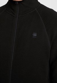 Chaqueta negra de cremallera con textura acanalada en los hombros, cierre con cremallera y un pequeño logo en relieve en el lado izquierdo del pecho.