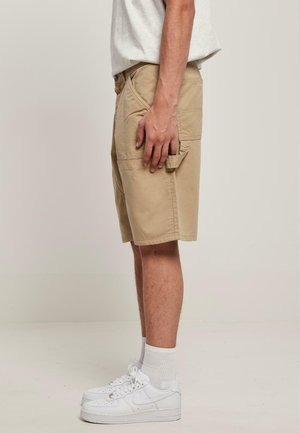 Beige Cargo-Shorts mit mehreren Seitentaschen, flacher Vorderseite und leicht umgeschlagenem Saum. Kombiniert mit weißen Sneakers und weißen Knöchelstrümpfen.