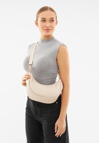 Borsa a mezzaluna beige realizzata in pelle liscia con accessori in tono oro e tracolla regolabile, tenuta da un modello con un top grigio.