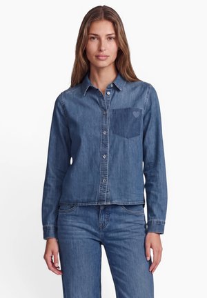Frau trägt ein mittelblaues Jeanshemd mit Knöpfen vorne und passende Jeans vor einem schlichten weißen Hintergrund.