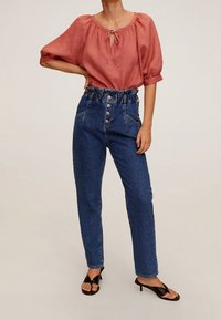 Blouse rouge à manches courtes avec un col noué ; jeans bleu délavé taille haute avec ceinture froncée et bouton sur le devant ; sandales noires.