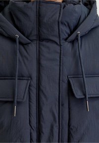 Veste matelassée bleu marine foncé avec col montant, deux poches avant à rabat et capuche avec cordons ornés d'embouts métalliques.