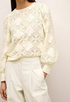 Femme portant un chemisier en dentelle crème à manches longues bouffantes et un pantalon crème taille haute cintré avec des plis.