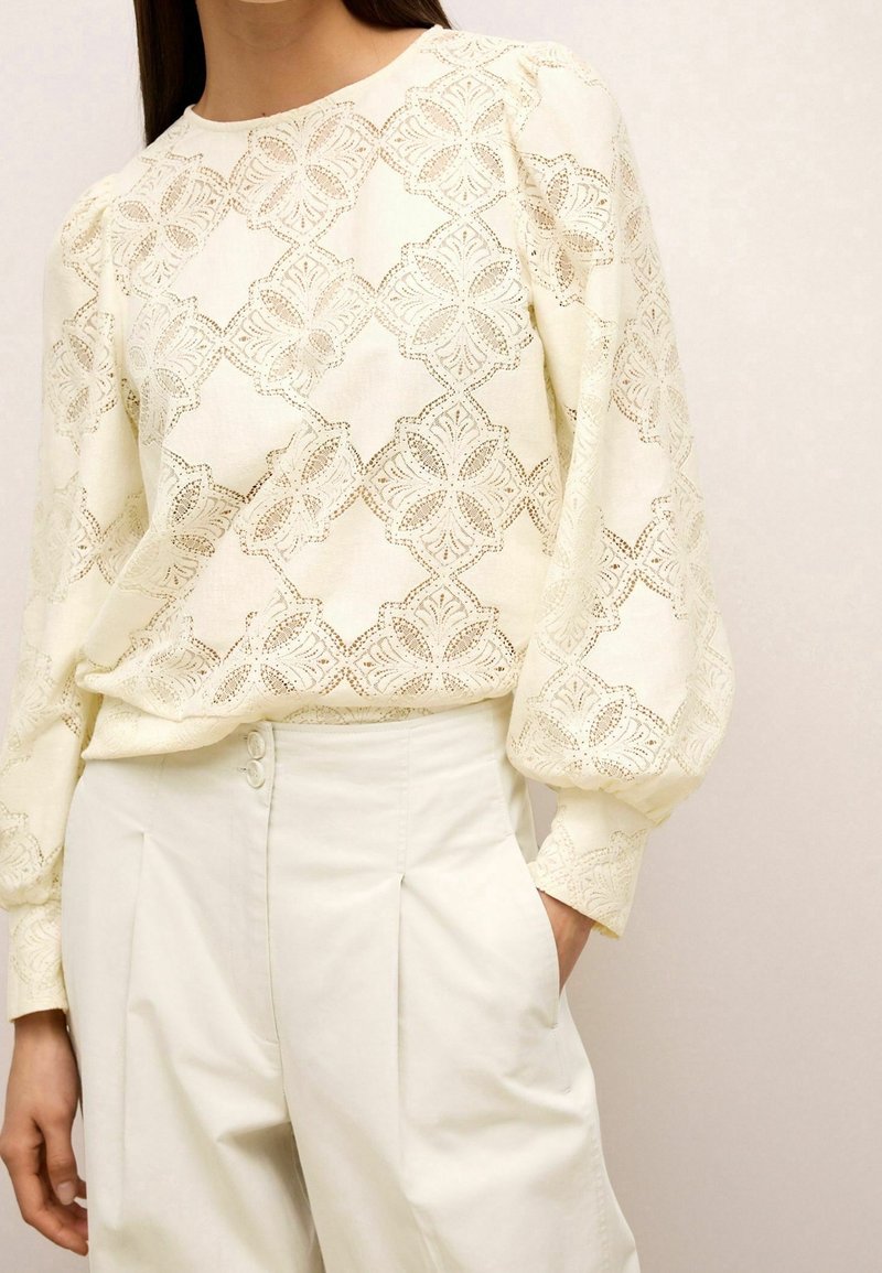 Femme portant un chemisier en dentelle crème à manches longues bouffantes et un pantalon crème taille haute cintré avec des plis.